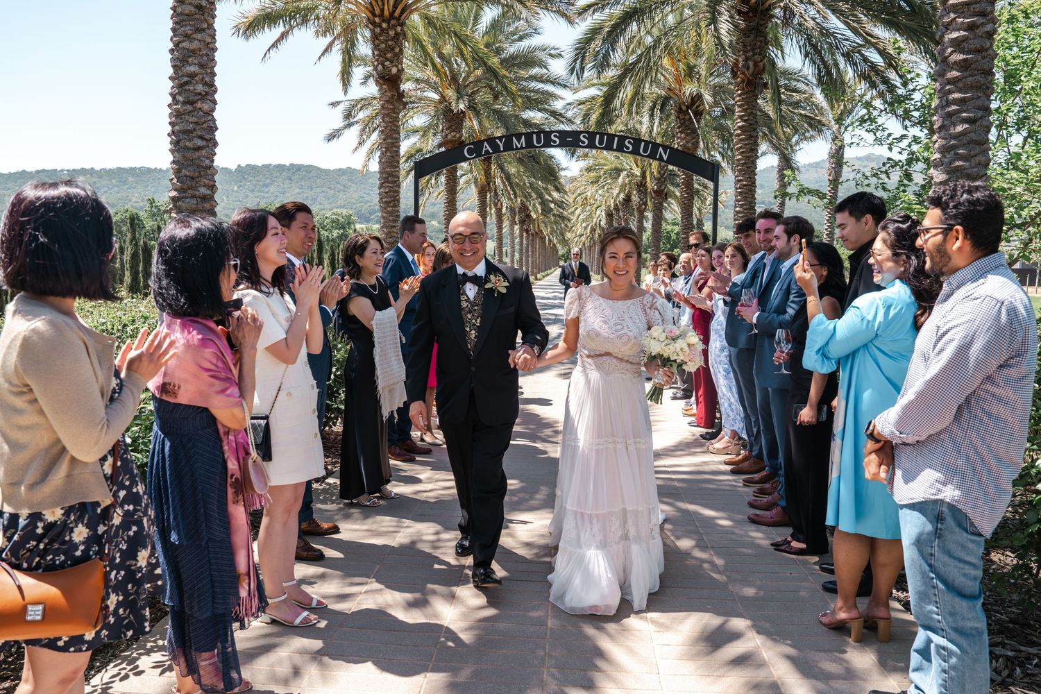 Bride and groom walk down outdoor wedding aisle lined with guests under palm trees and decorative arch.