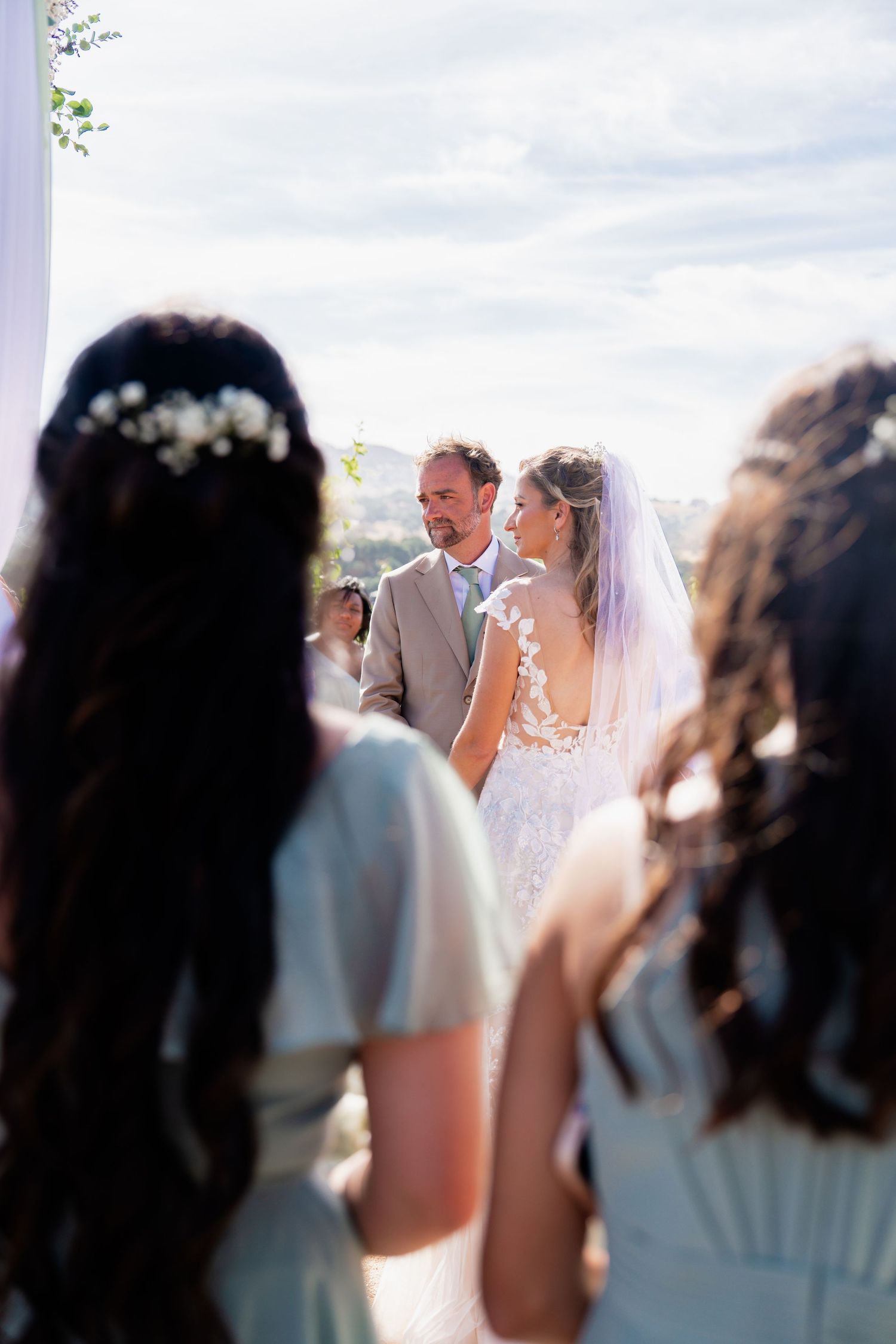 Bride and groom exchange vows outdoors as bridesmaids with floral hair accessories look on at elegant wedding ceremony.
