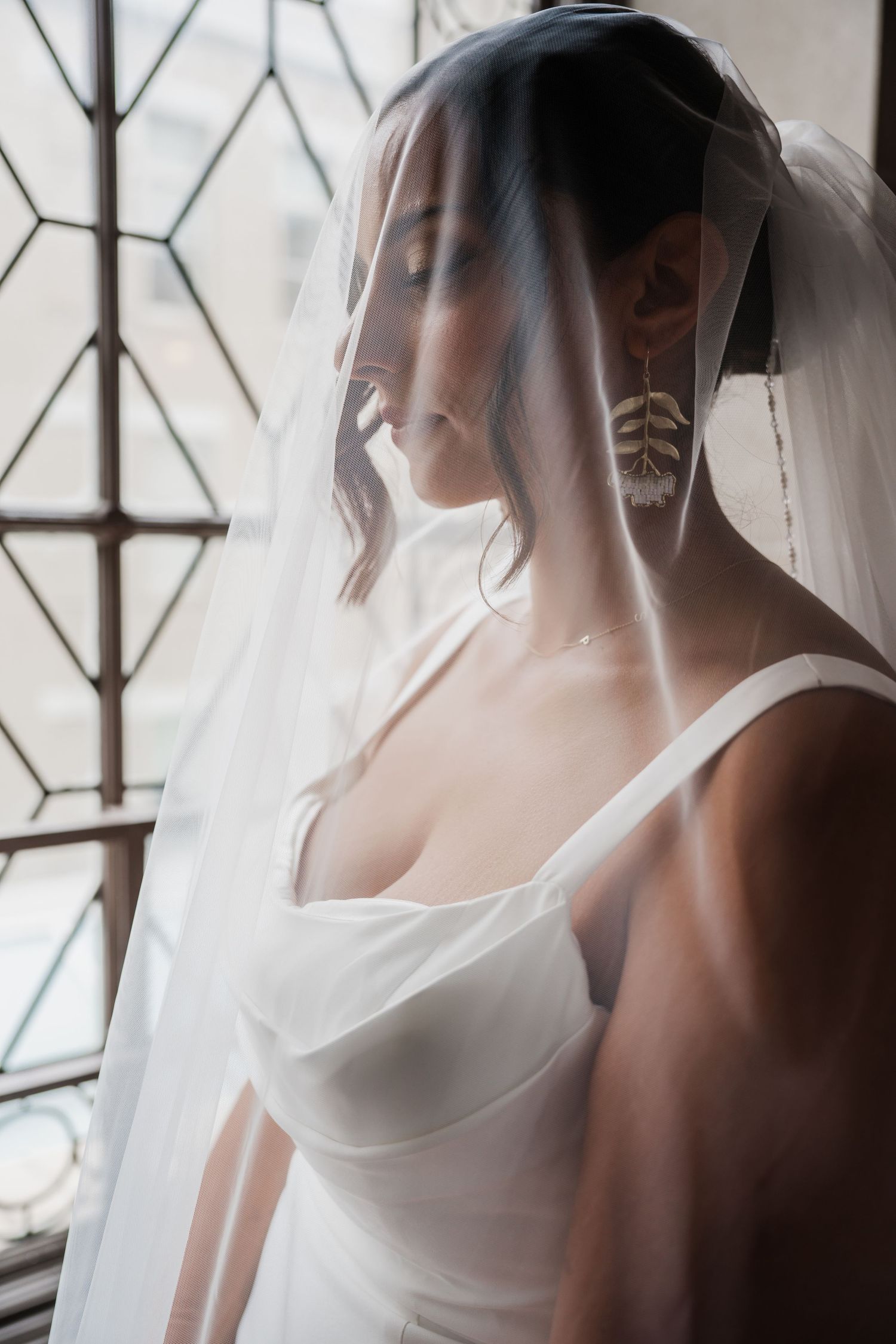 Bride in white gown and veil with feather earrings poses by geometric window in soft natural light.