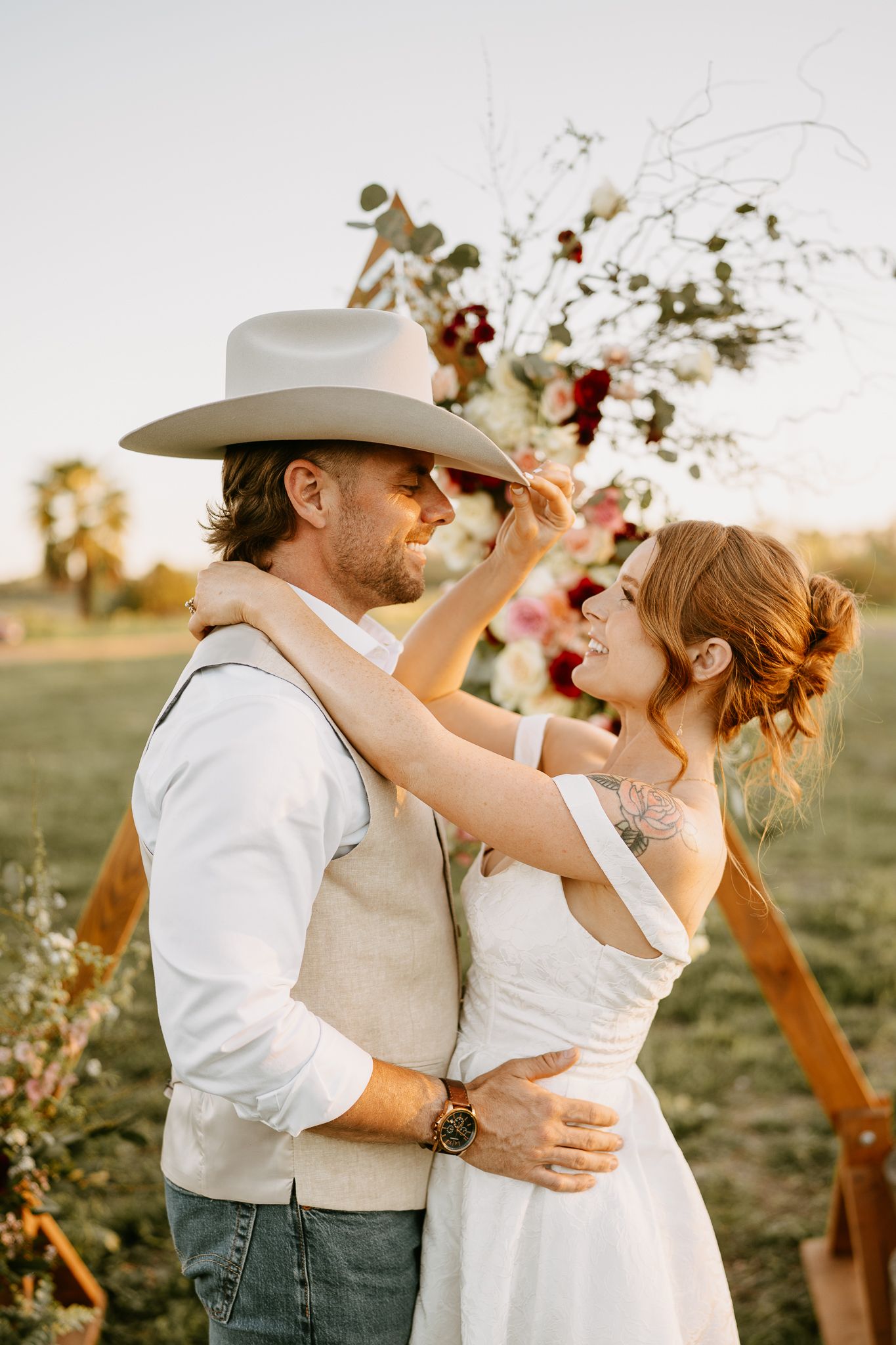 Bride and groom embrace outdoors at golden hour, she tips his cowboy hat while smiling, floral arch backdrop.