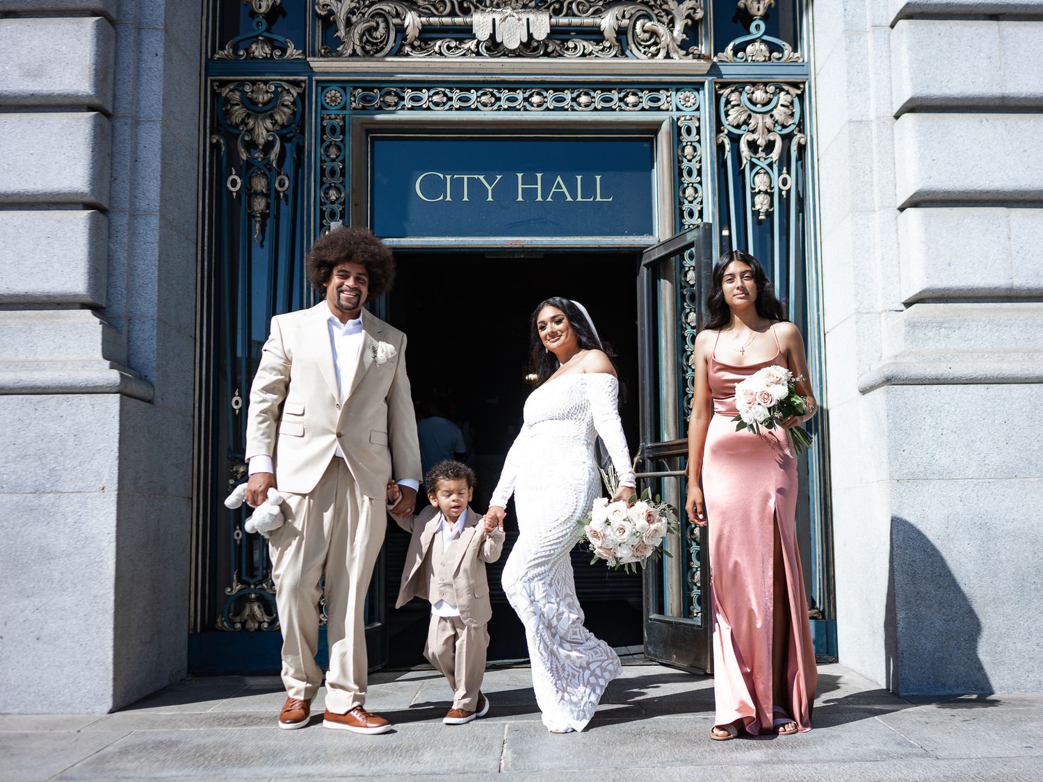 Wedding party exiting City Hall with bride in white lace gown and bridesmaid in pink dress.
