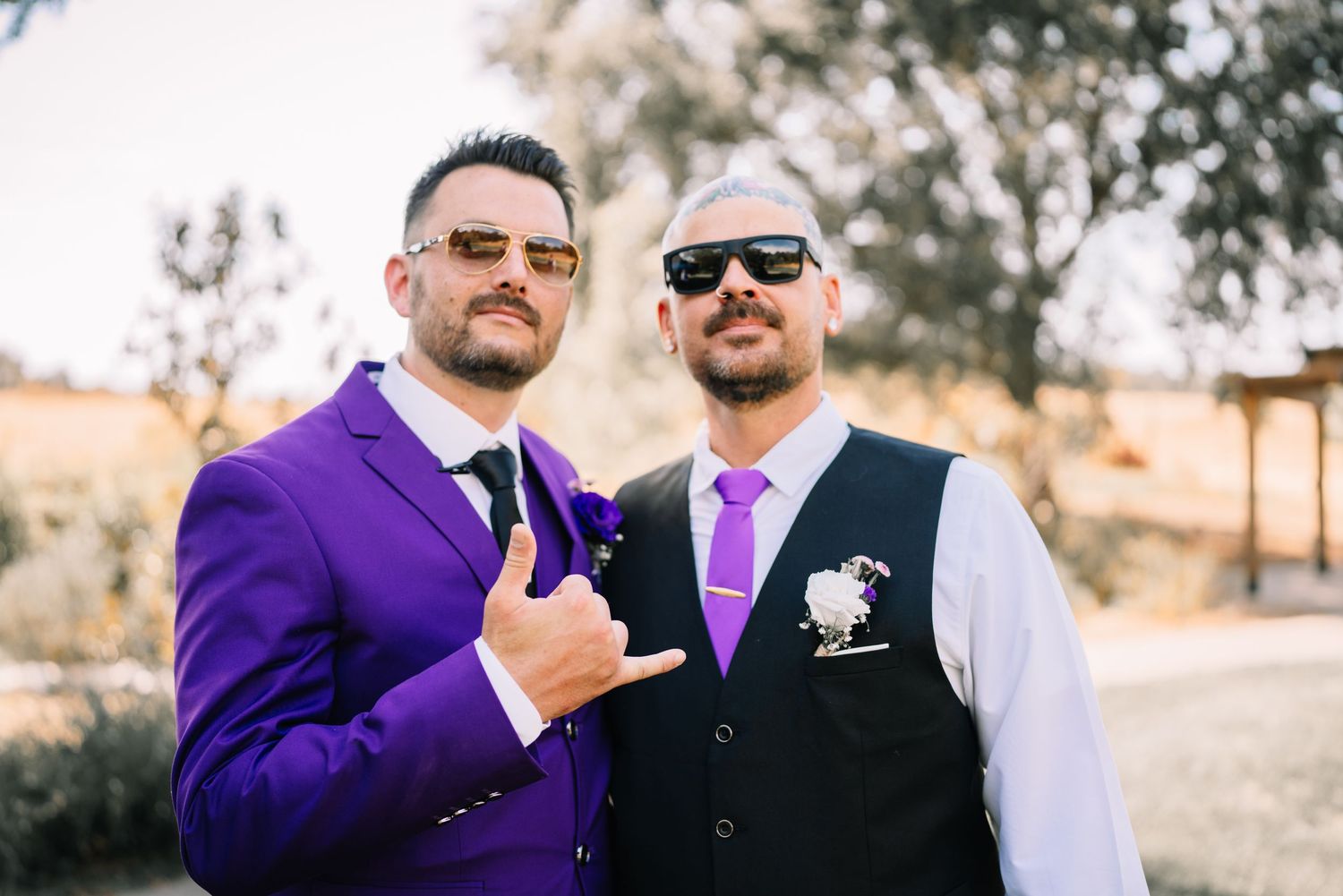 Two men in purple suits and sunglasses posing at a wedding, one giving a shaka sign outdoors.