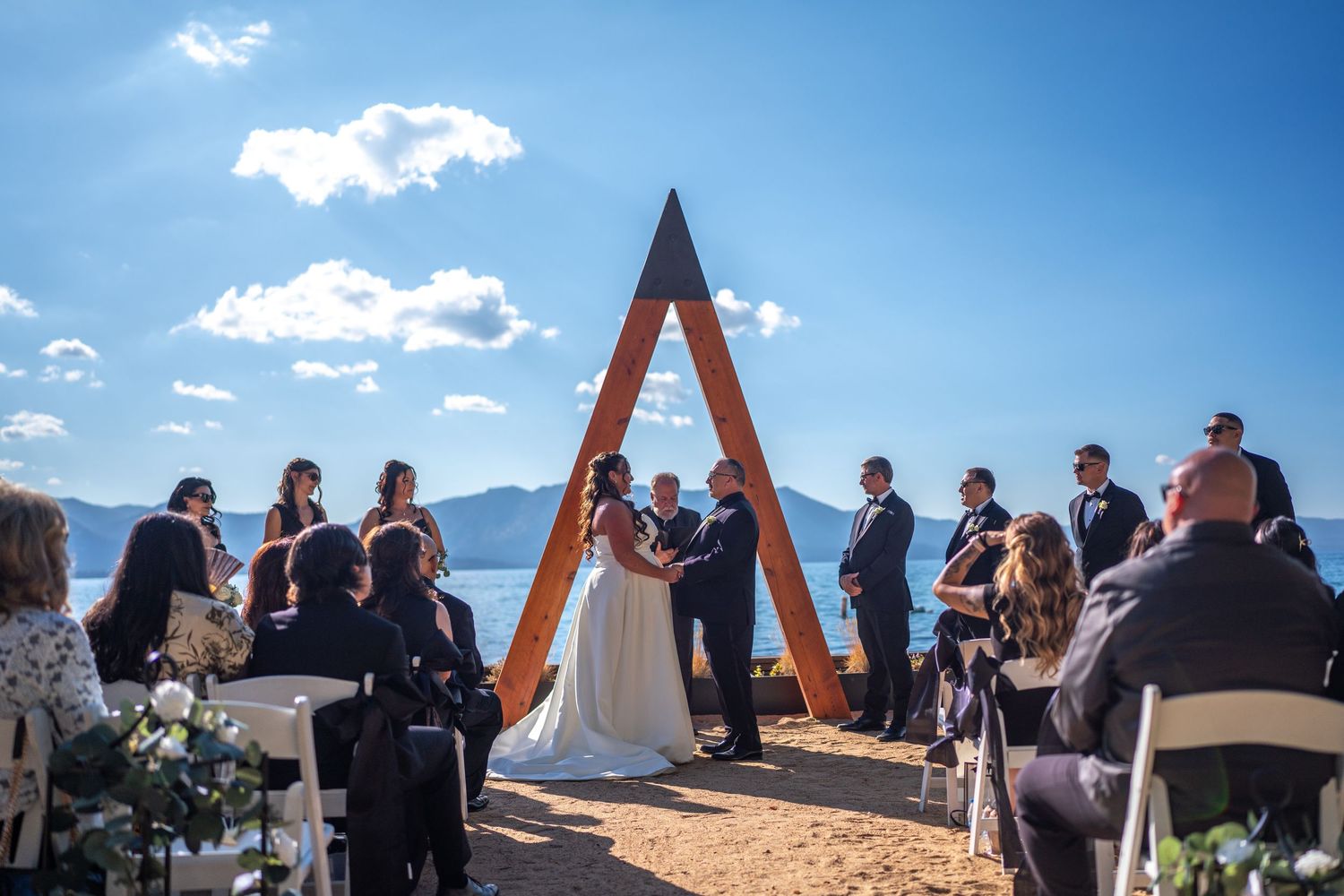 Outdoor wedding ceremony under a wooden A-frame arch with mountain views and blue sky backdrop.