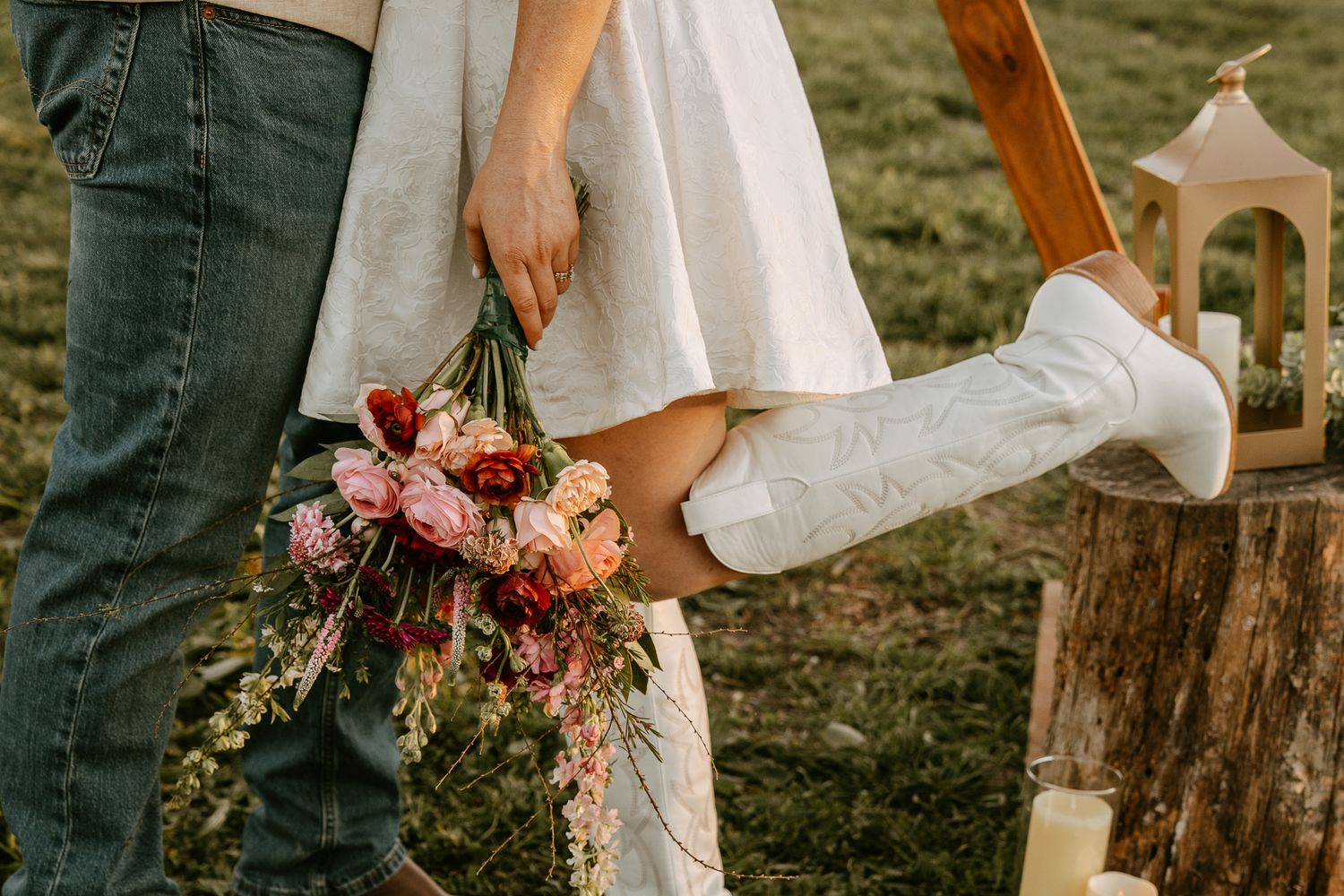 Bride in white dress and cowboy boots holds floral bouquet beside groom in jeans at outdoor wedding.