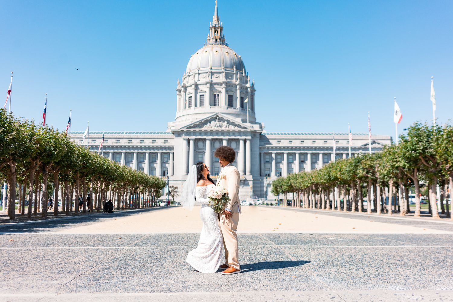 Newlywed couple poses in front of San Francisco City Hall on a sunny day, surrounded by tree-lined plaza.