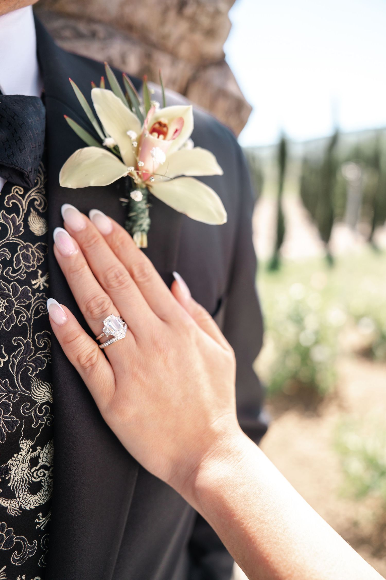Close-up of a hand wearing an engagement ring with a delicate orchid boutonniere.