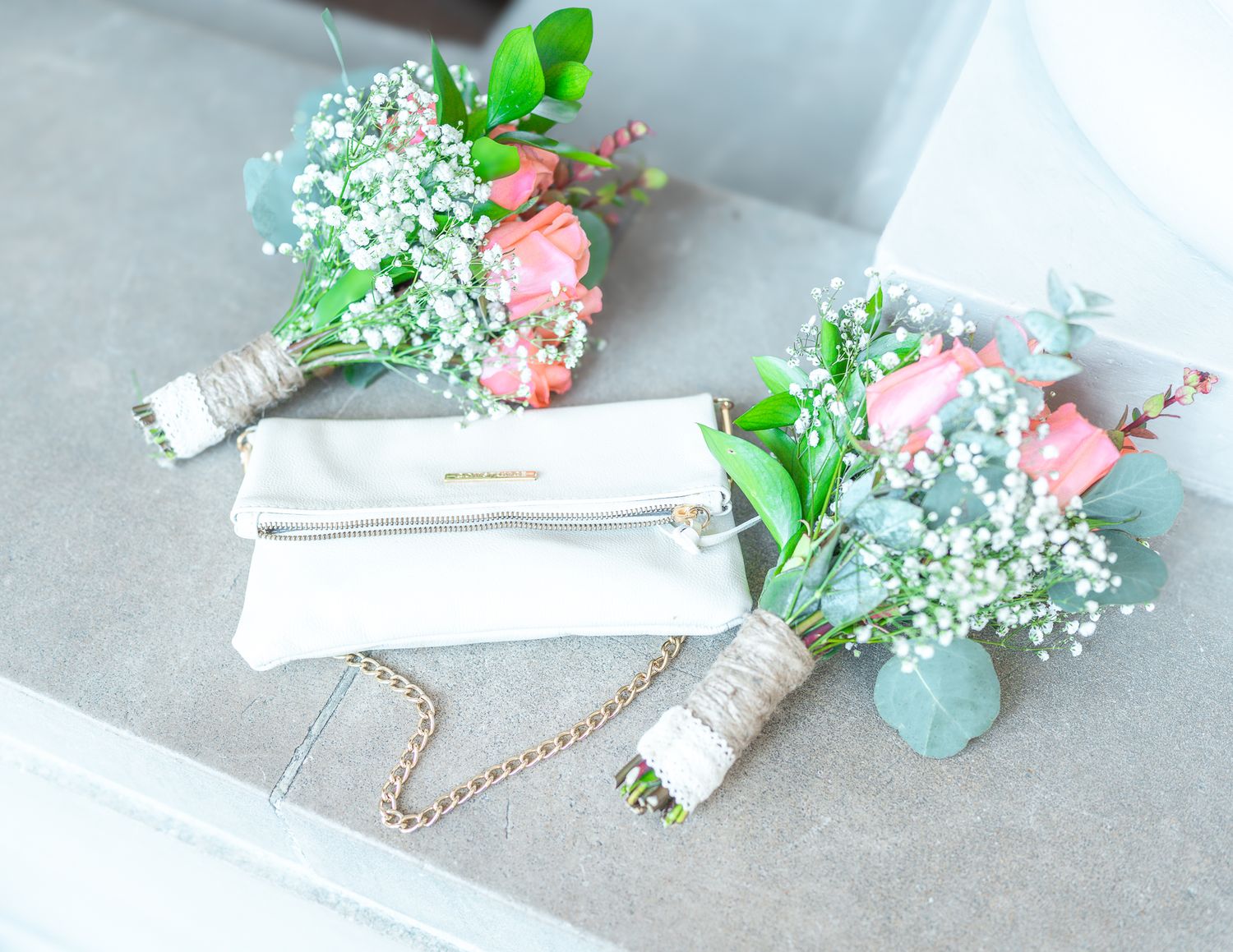 Two pink rose and baby's breath wedding bouquets beside a white clutch purse on gray surface.