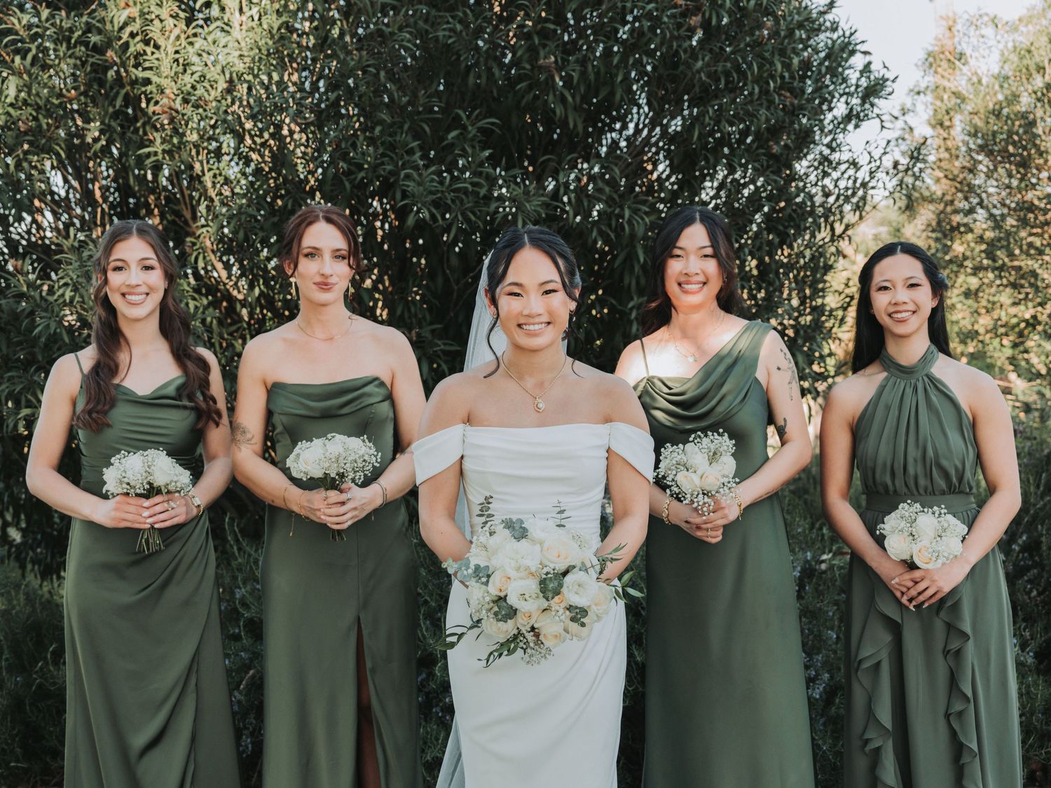 Bride in white gown with veil surrounded by four bridesmaids in olive green dresses holding white bouquets outdoors.