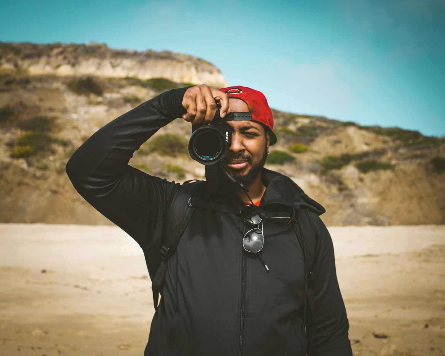 A man in a red beanie and black jacket photographs with a DSLR camera on a sandy beach with rocky cliffs.