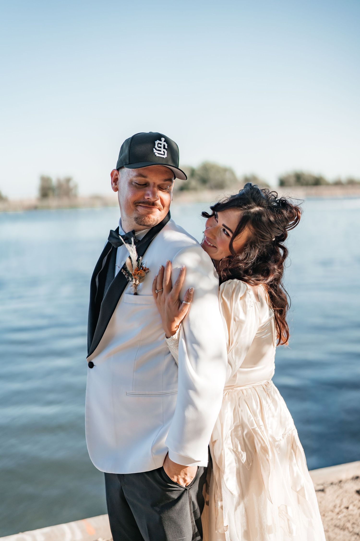 Couple posing by a lake on their wedding day, groom in white blazer and cap, bride in lace dress embracing him.