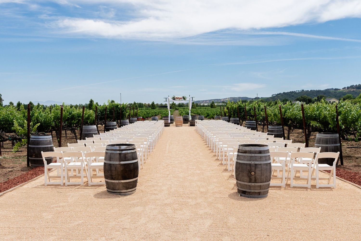 Wedding ceremony site set up between vineyard rows with wine barrels and white chairs.