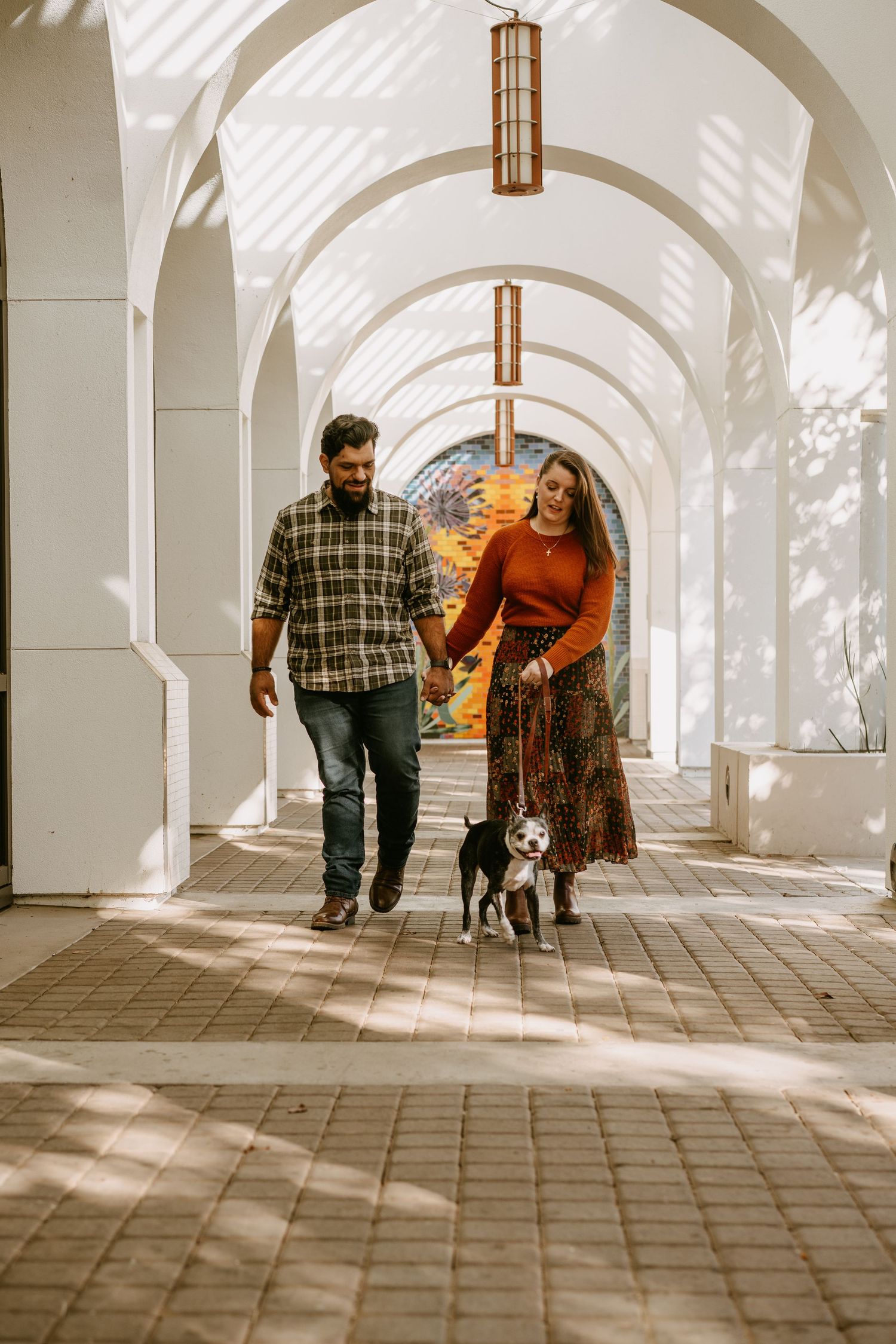 A couple walking with their small dog under arched walkway with dramatic shadows and lighting.