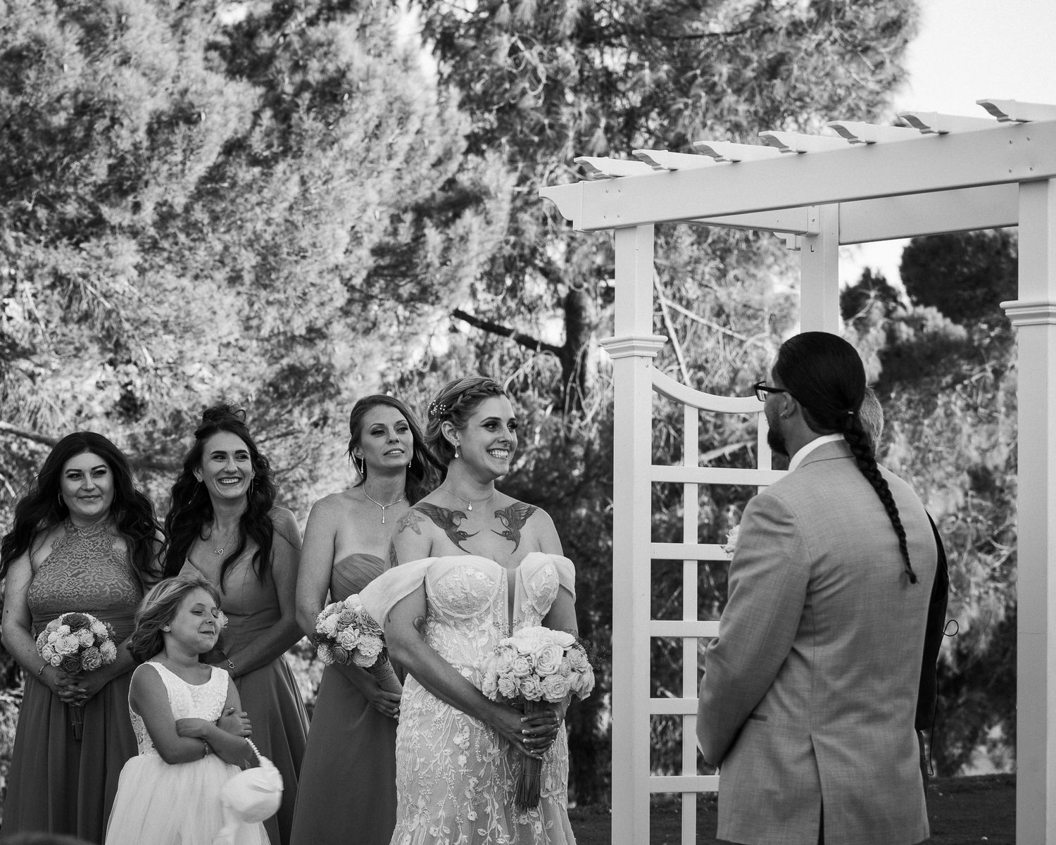 Black and white photo of bride smiling at groom during outdoor wedding ceremony with bridesmaids.
