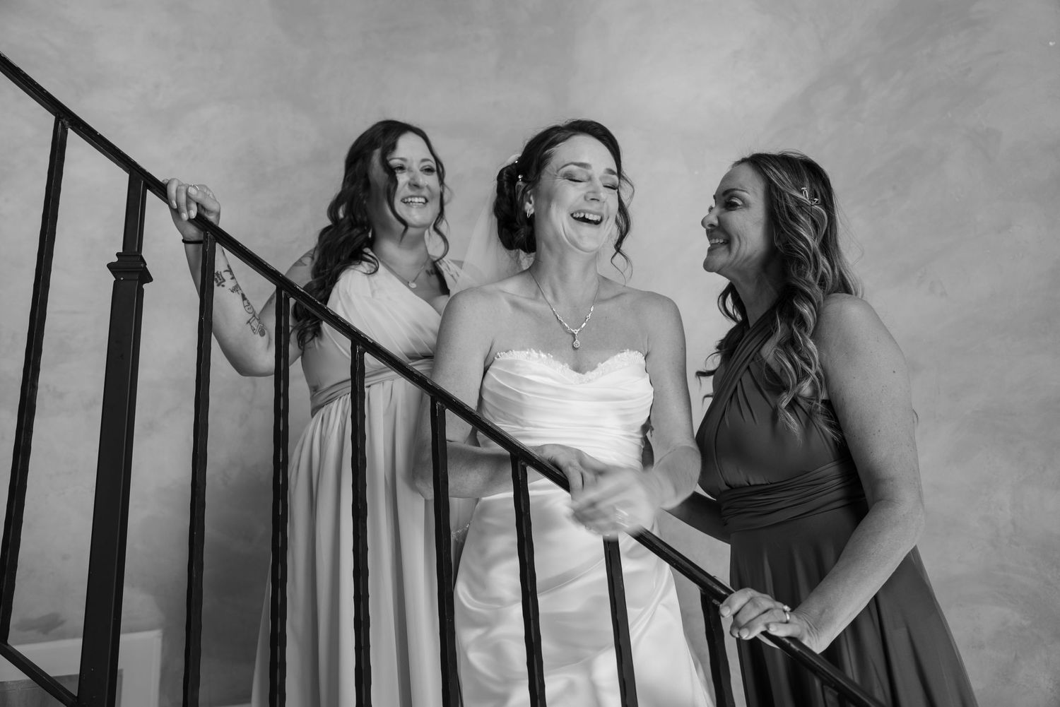 Black and white photo of a bride laughing with two bridesmaids on a staircase during wedding preparations.