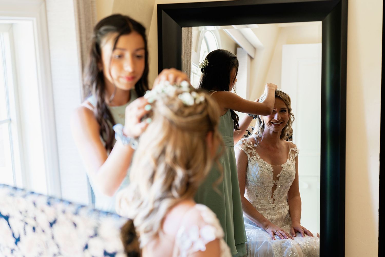 Bridesmaid adjusting bride's floral hair accessory reflected in elegant black-framed mirror on wedding day.