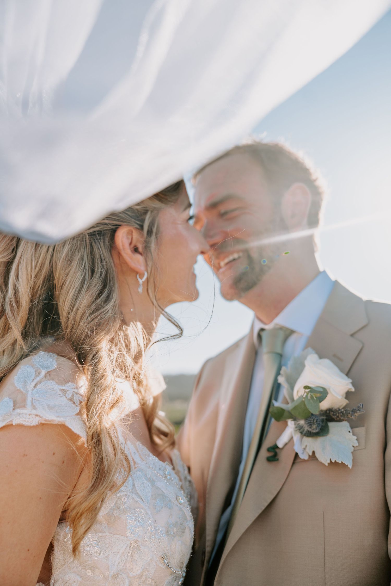 Bride and groom sharing a romantic moment under a flowing veil, backlit by golden sunlight outdoors.