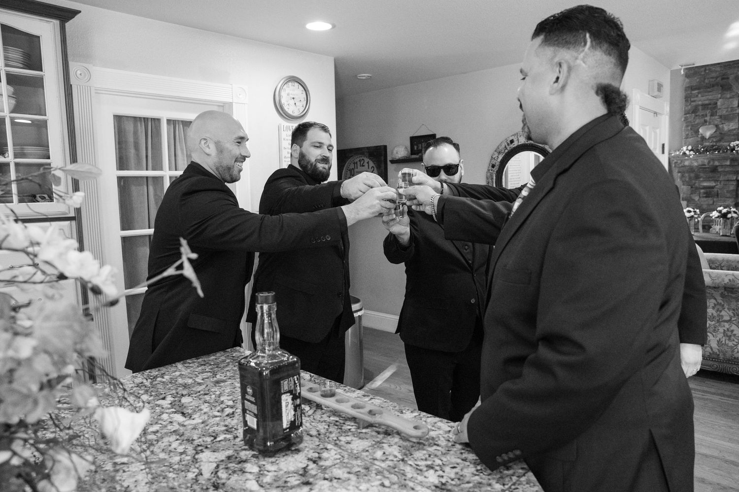 Groomsmen in black suits toasting with whiskey at a kitchen counter in a black and white wedding photo.