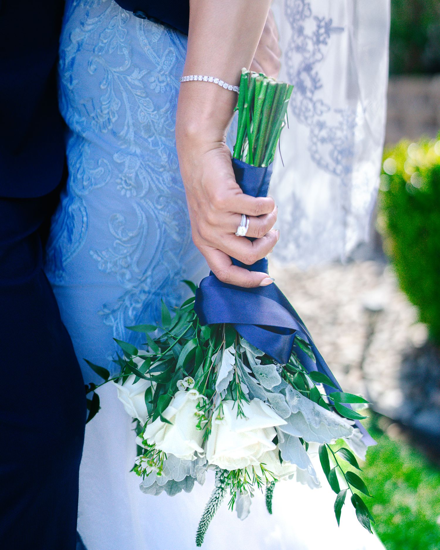 Bride holding elegant white rose and greenery bouquet tied with navy ribbon at outdoor wedding.
