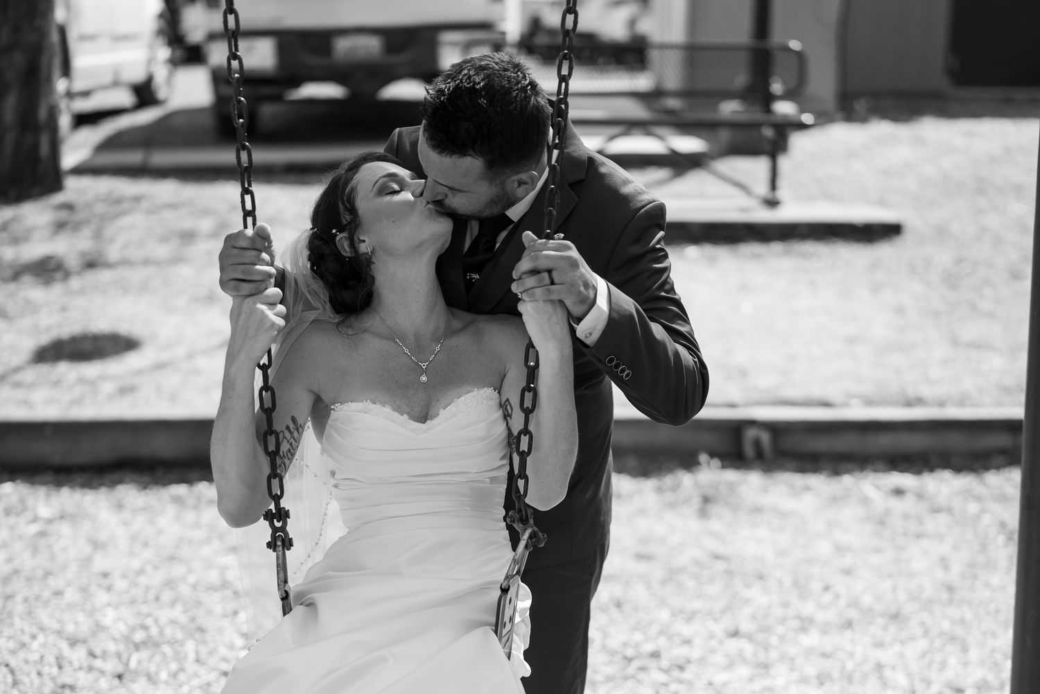 Black and white photo of a bride on a swing being kissed by her groom in a romantic outdoor wedding moment.