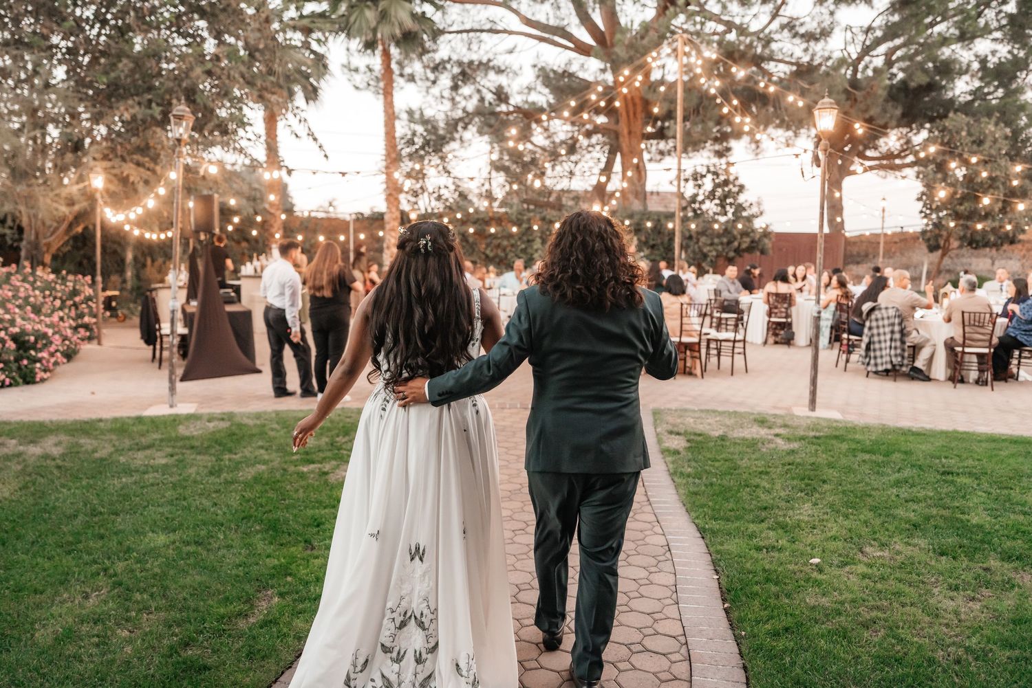 Bride in a white embroidered gown and groom in a dark suit walk arm in arm along a brick path toward an outdoor wedding reception at dusk.