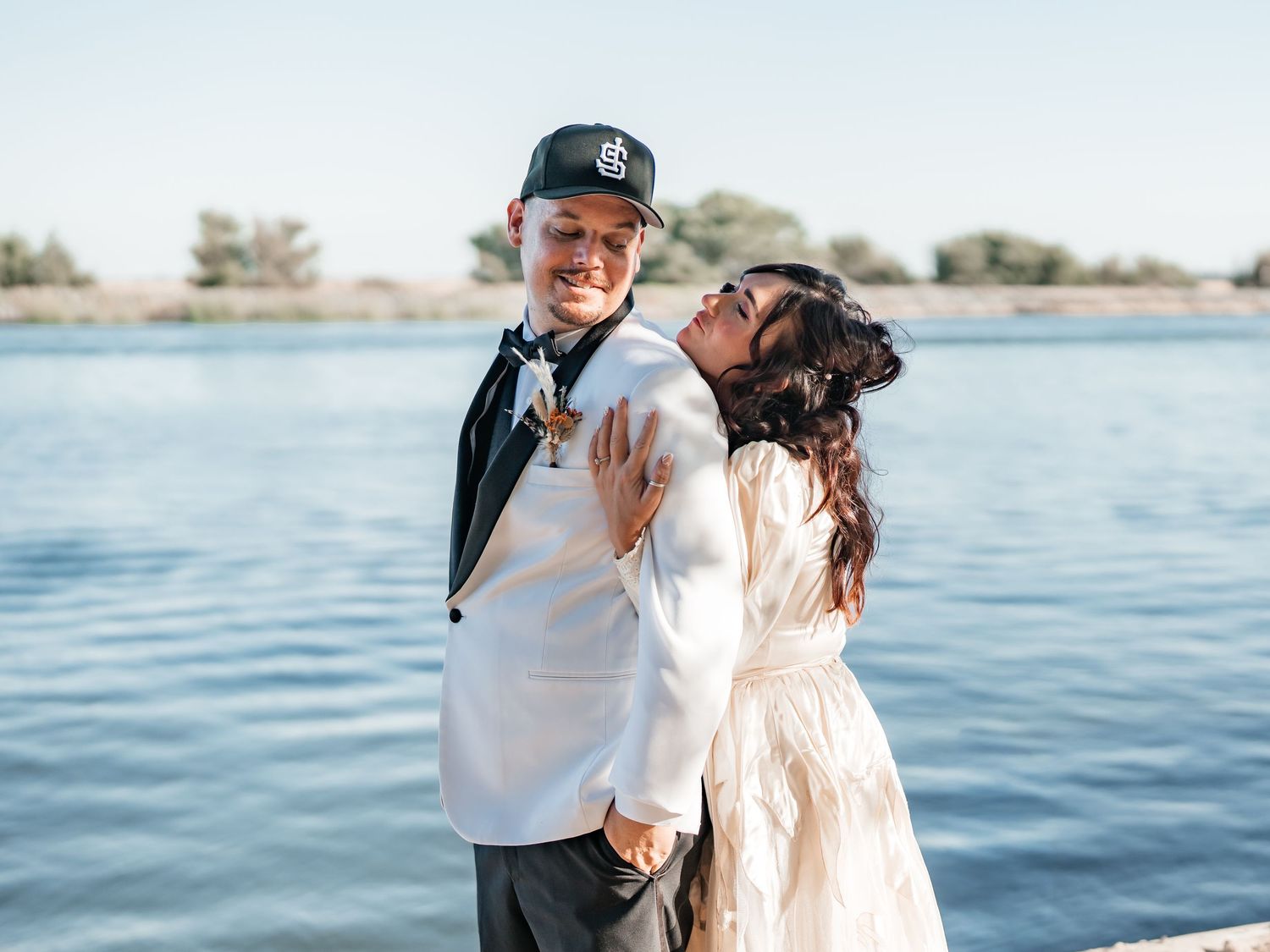 Couple in wedding attire embracing by a lake, groom in white jacket and cap, bride in white dress kissing his cheek.