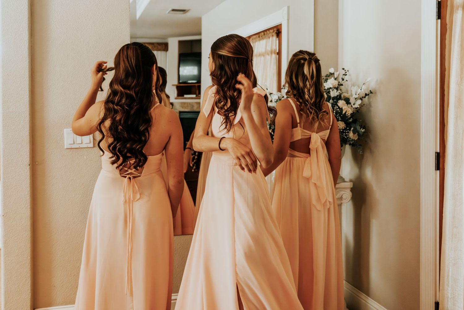 Bridesmaids in blush pink gowns stand before a mirror helping bride with her wedding dress during bridal preparations.