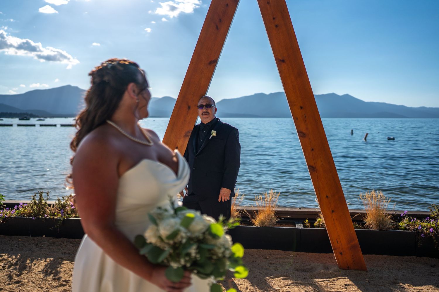 Bride holding white bouquet stands before groom at lakeside wedding ceremony with wooden arch and mountain backdrop.