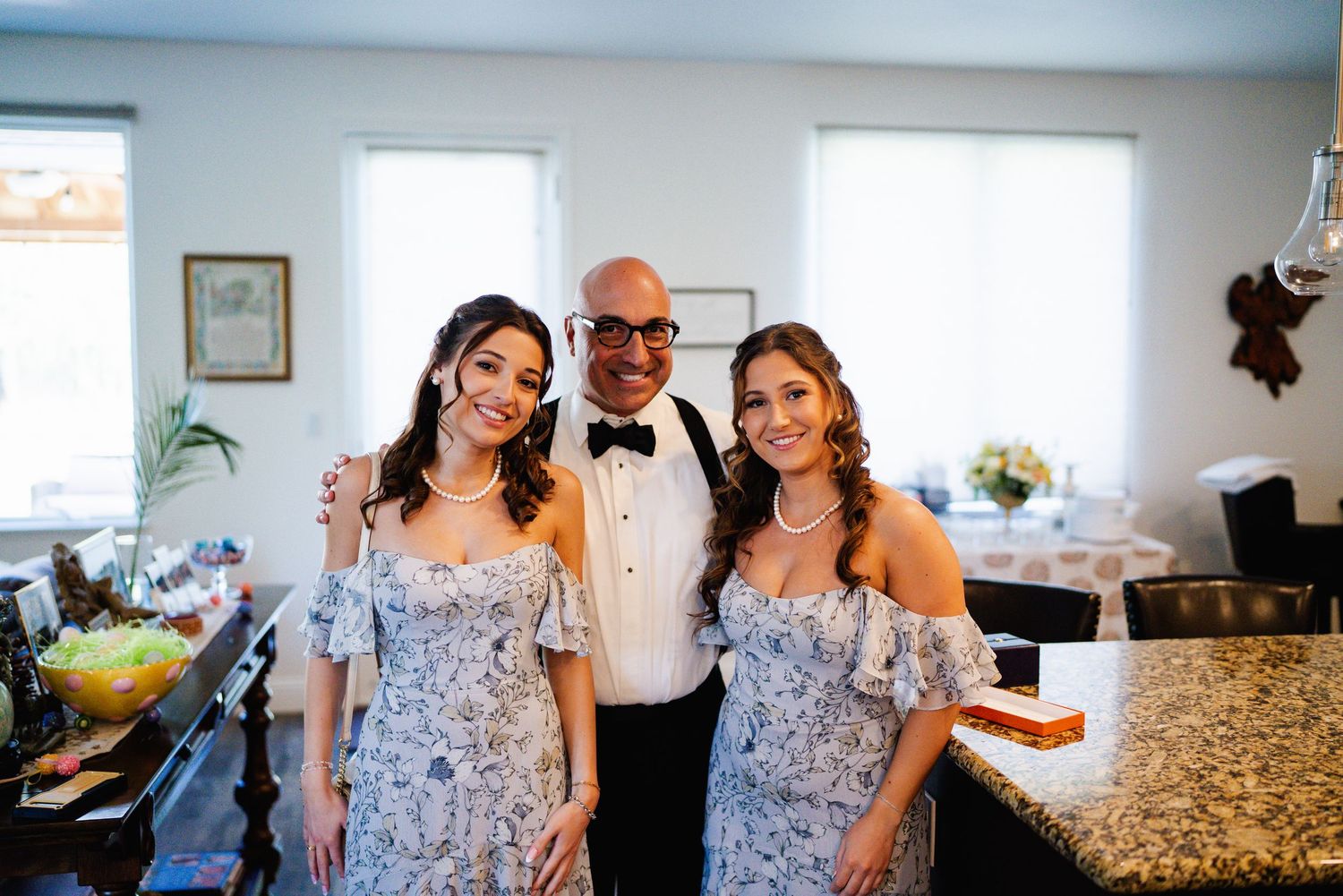 Man in tuxedo poses with two women in matching floral dresses at an elegant indoor celebration.