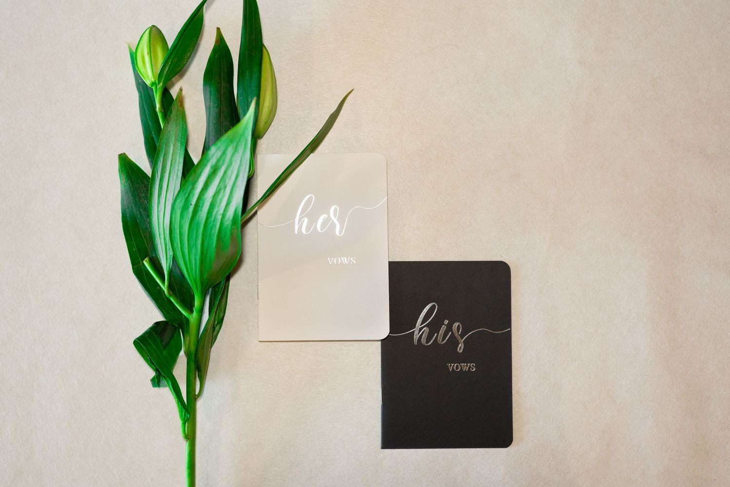 His and hers wedding vow books in black and white with calligraphy script, styled with tropical green leaves.