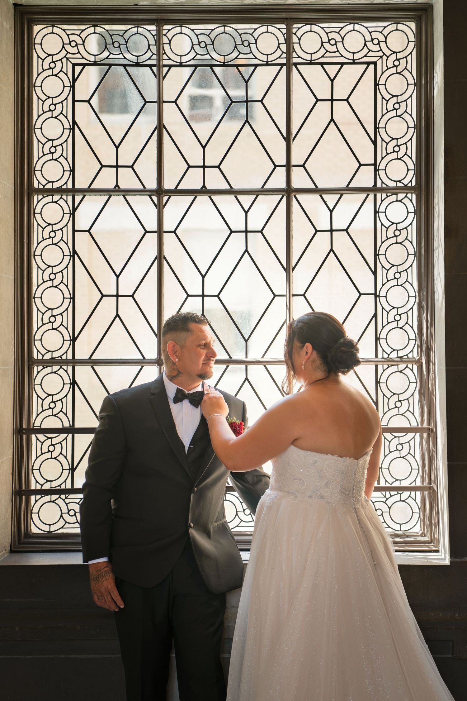 Bride adjusting groom's boutonniere in front of ornate stained glass window in elegant wedding venue.