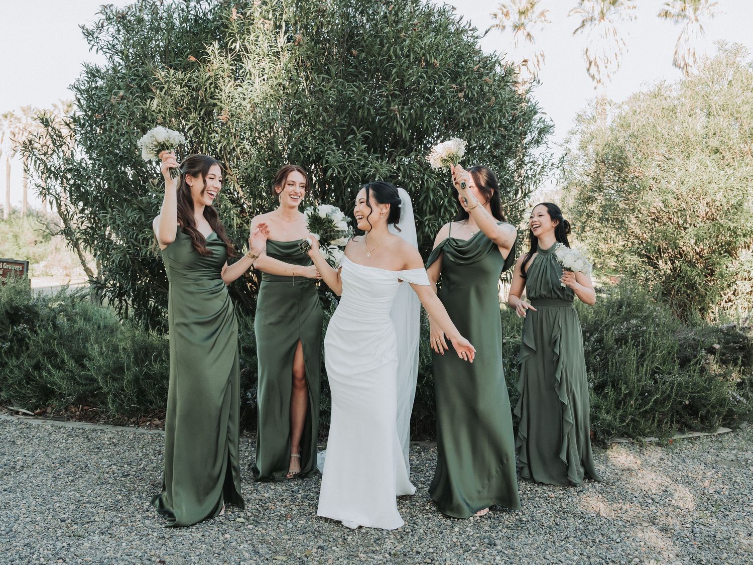 A bride in a white gown laughs with four bridesmaids in olive green dresses, all holding white bouquets outdoors among lush green trees.