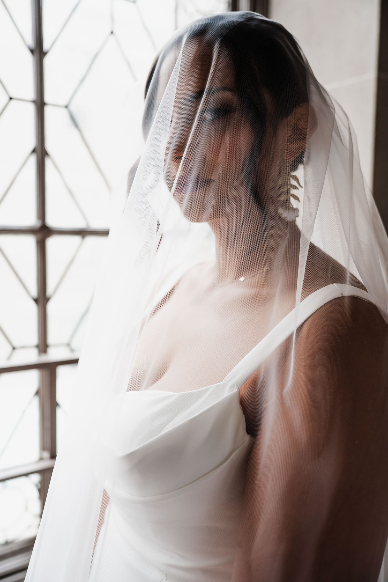 Bride in white gown with veil draped over face, standing by ornate window in soft natural light.