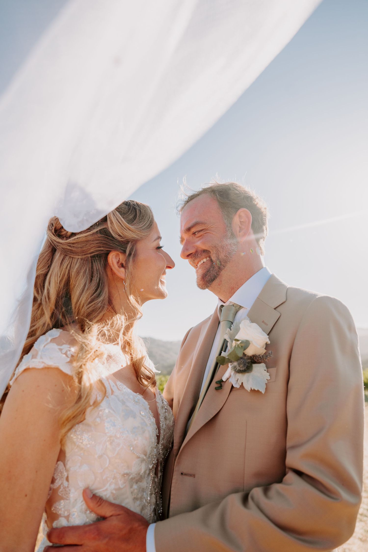 Bride and groom smiling at each other under flowing veil in golden sunlight, groom in tan suit with white boutonniere.