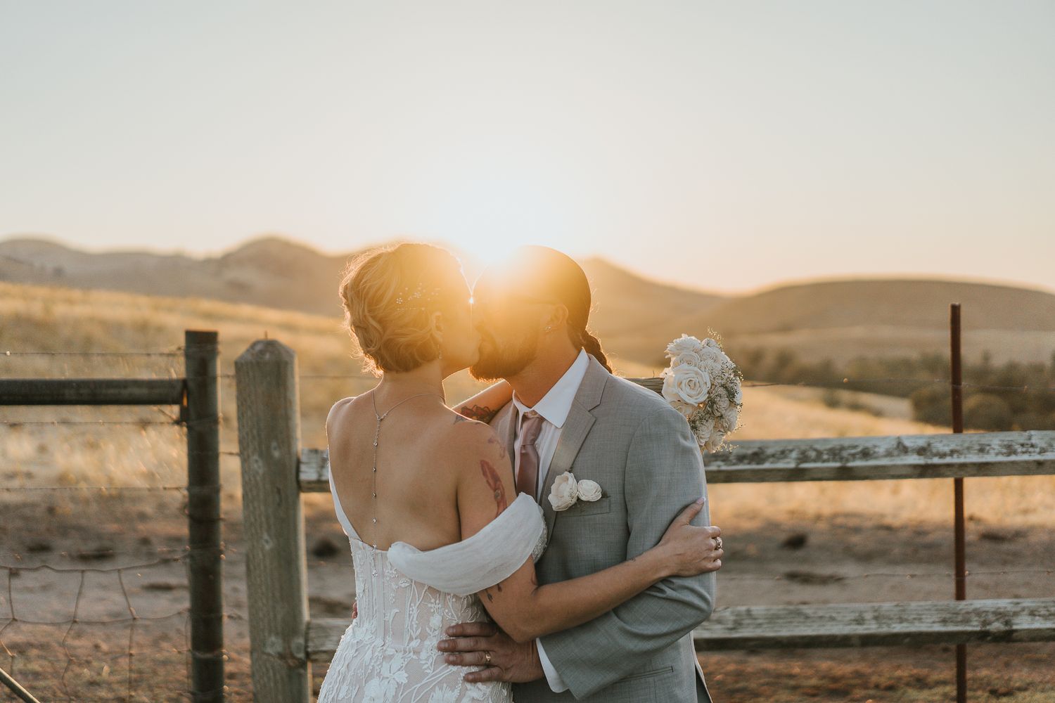 A bride and groom share a romantic kiss at golden sunset by a rustic fence in an outdoor wedding.
