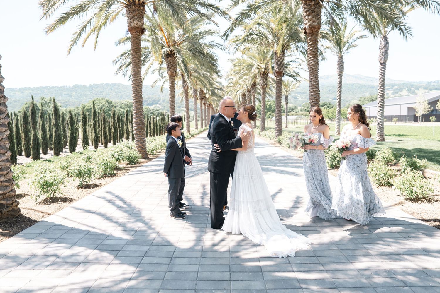 Wedding ceremony taking place on an elegant palm-lined pathway with mountains in the background.