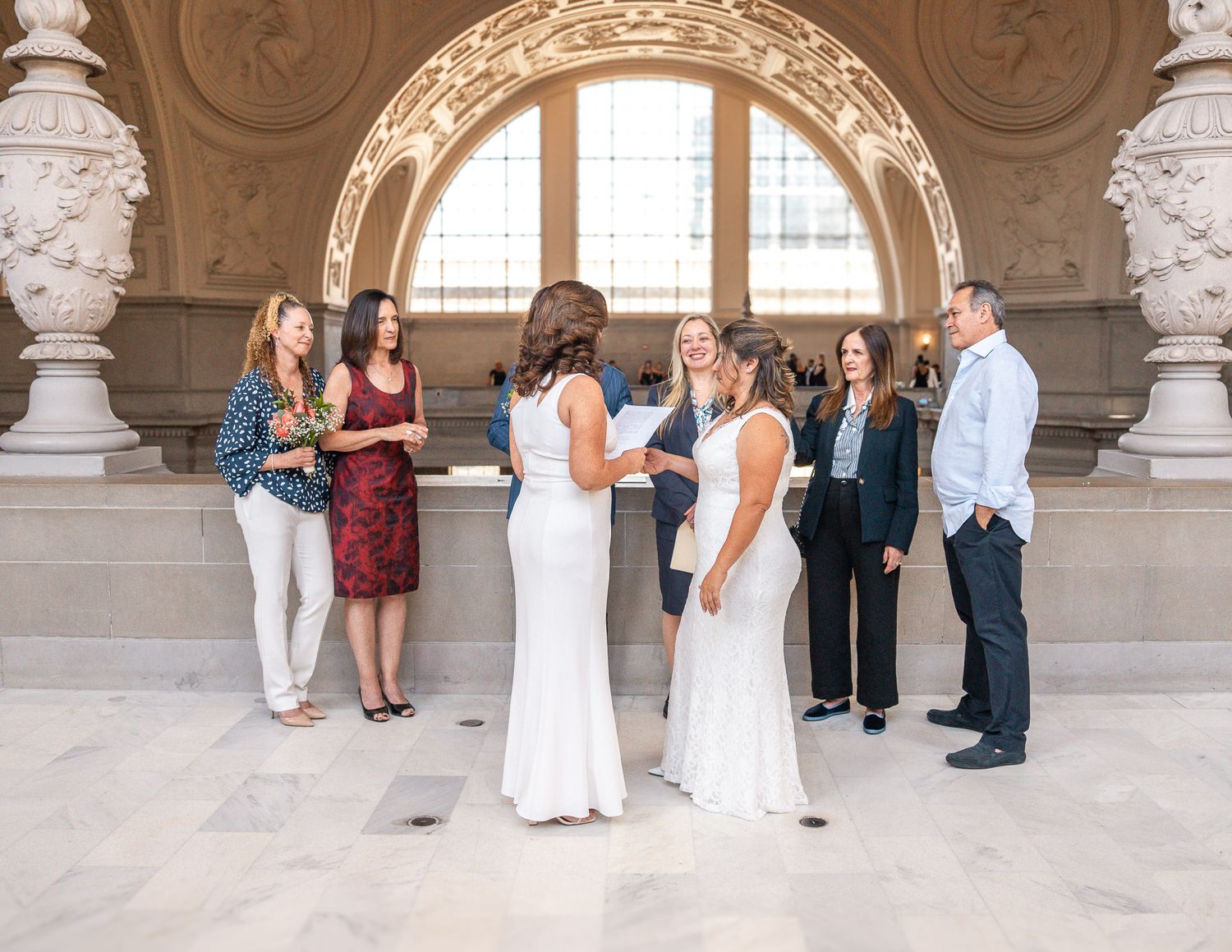Two brides in white dresses exchange vows surrounded by guests inside a grand ornate building.