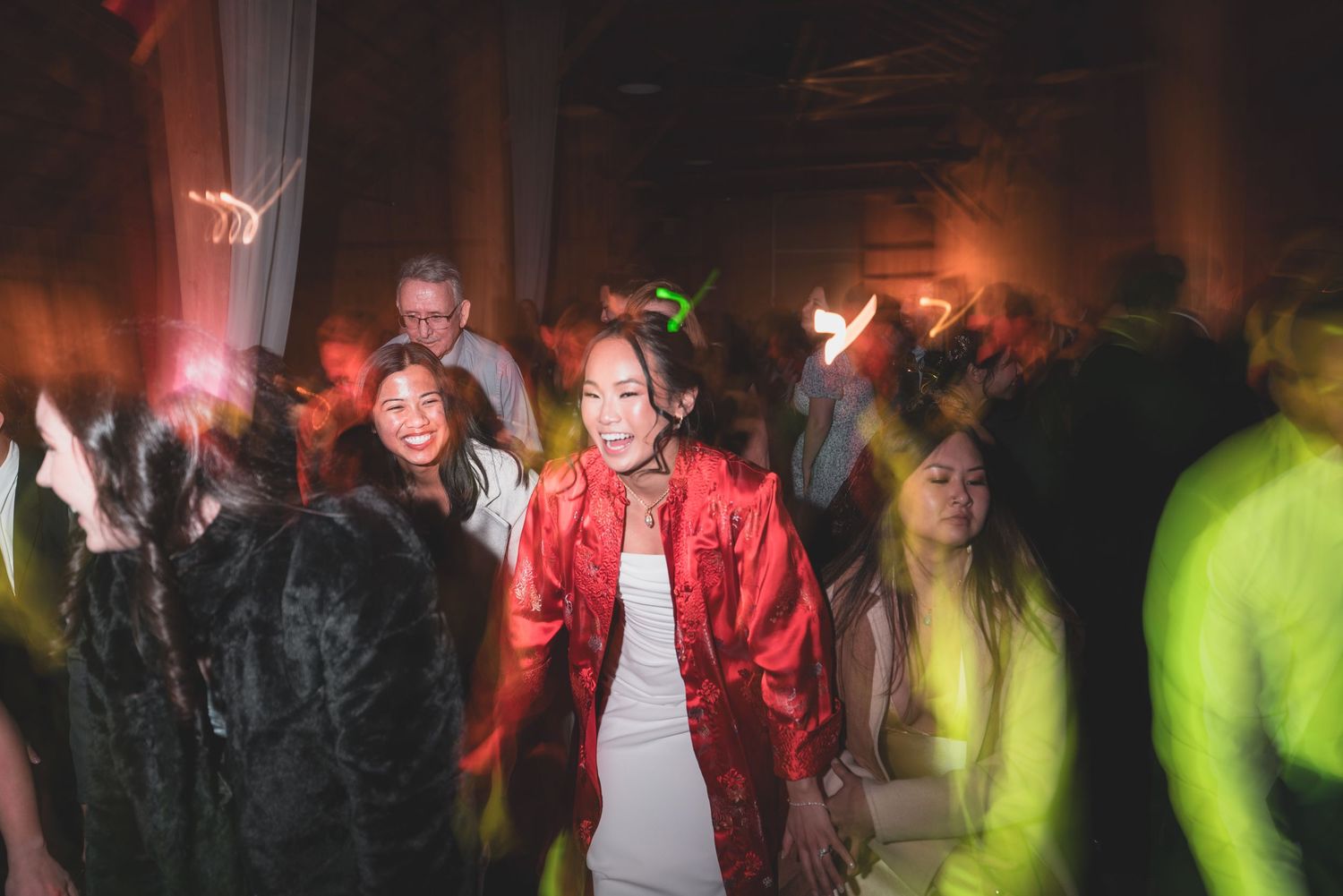 People dancing at a lively party, woman in red jacket and white dress stands out in the crowd.