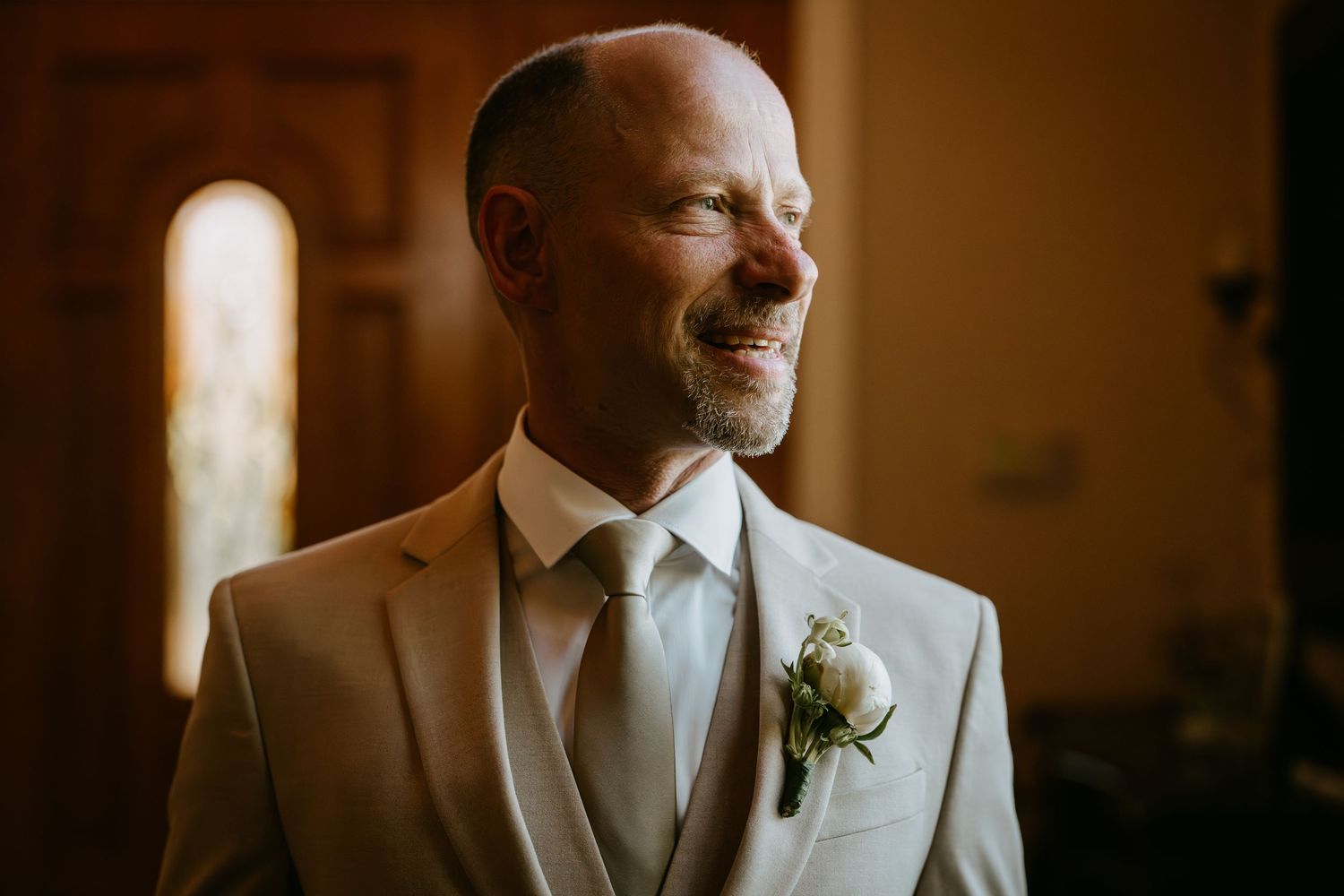 Smiling groom in a beige suit with boutonniere stands in warm light, radiating joy on his wedding day.