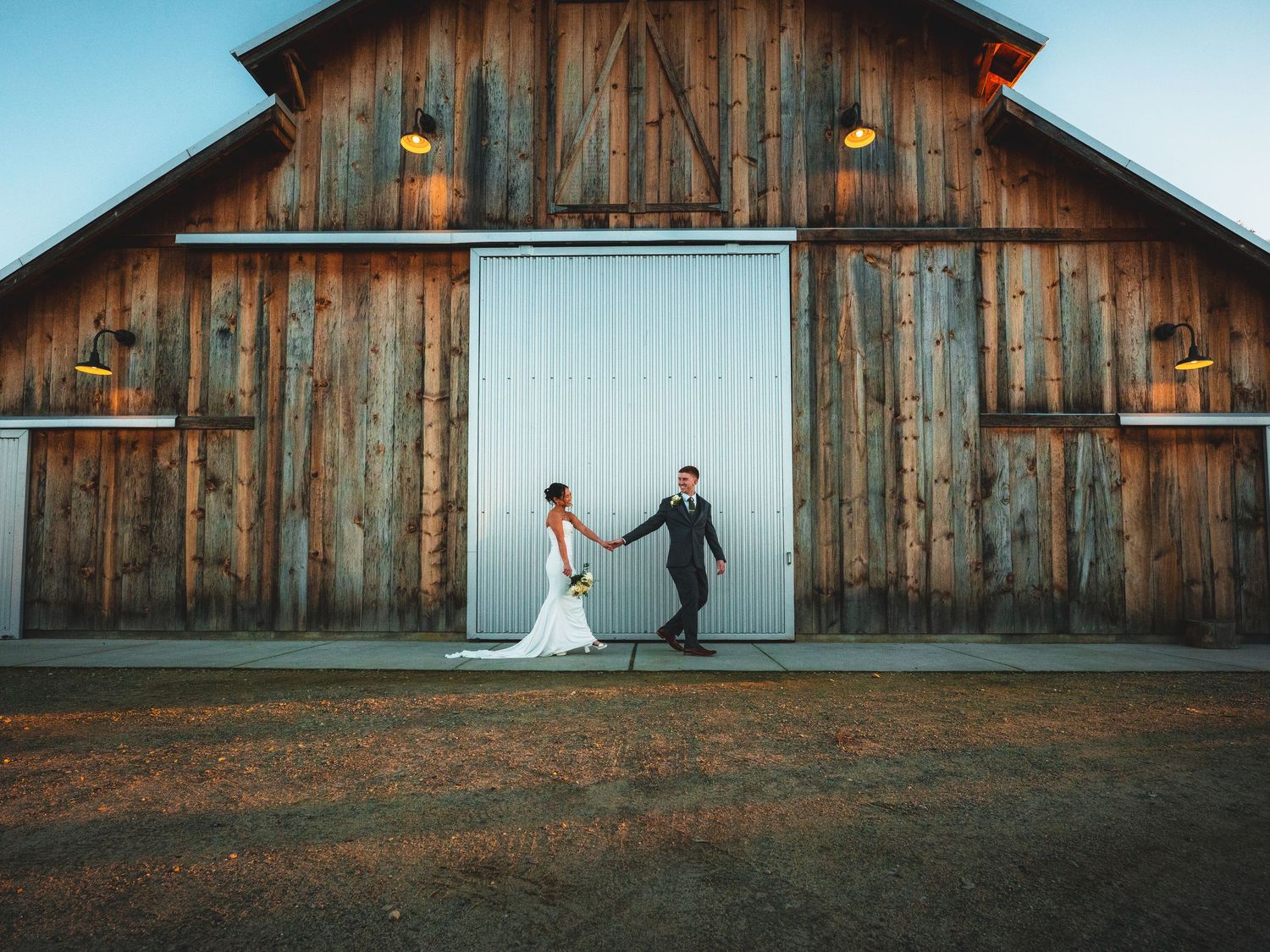Bride and groom hold hands outside a rustic wooden barn venue at dusk on their wedding day.