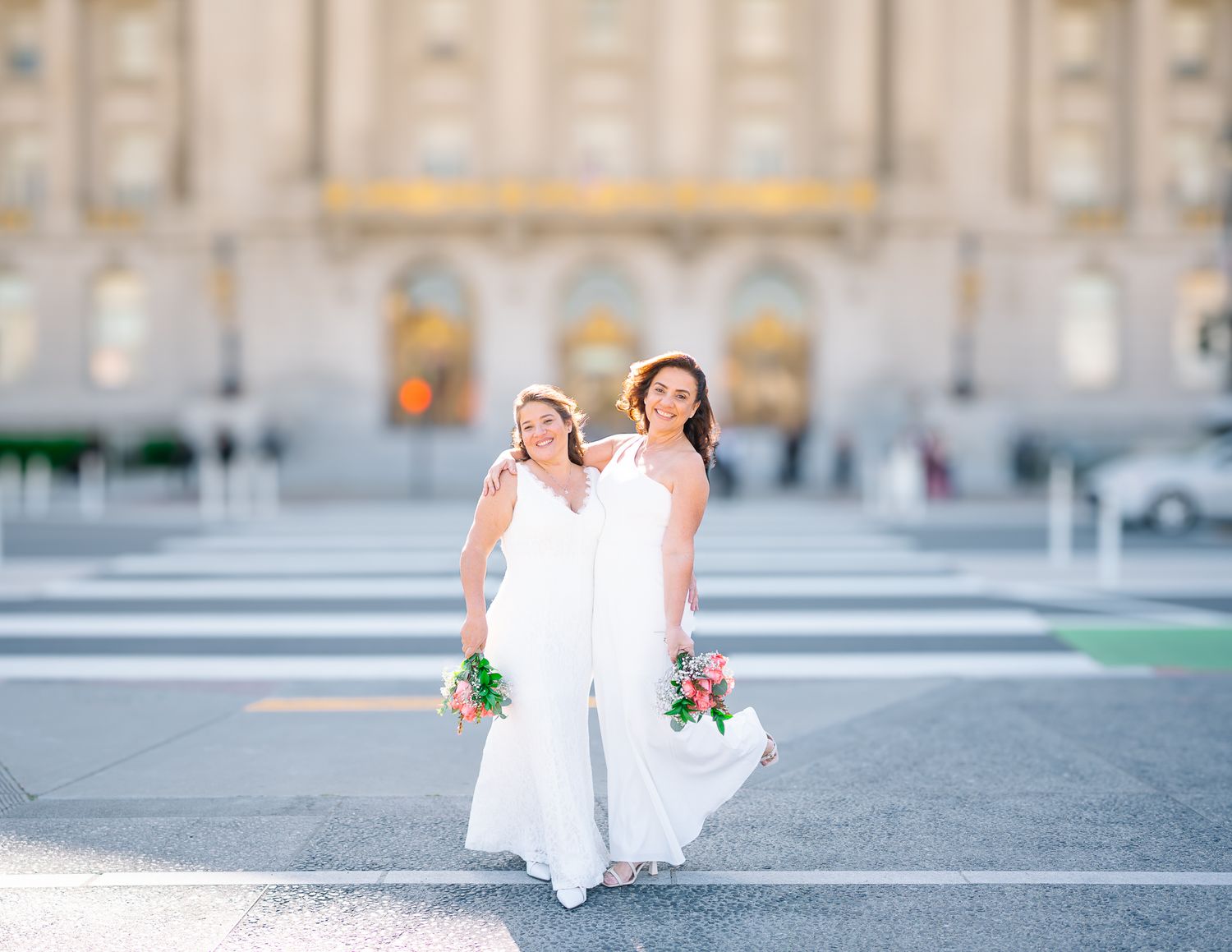 Two brides in white dresses hold colorful bouquets while crossing street in front of grand building.