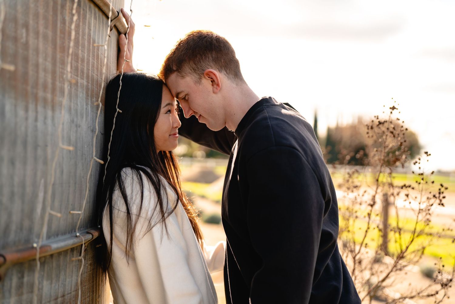 Romantic couple embraces against rustic barn wall in warm sunset lighting.