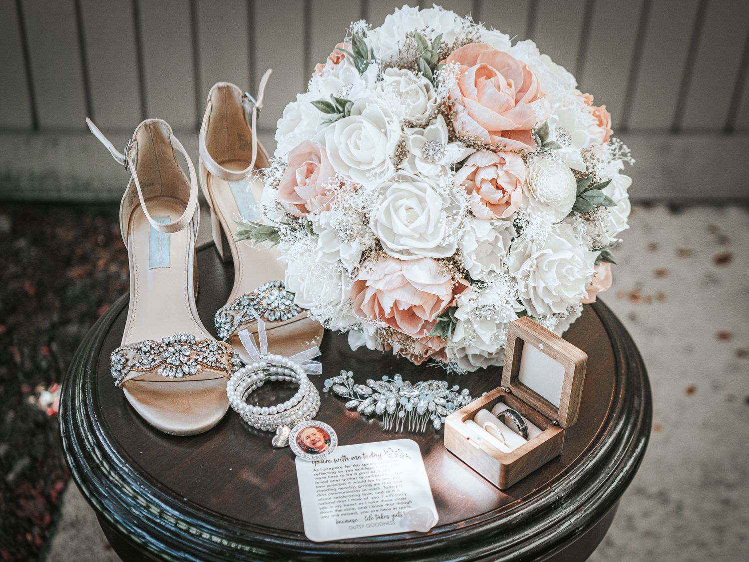 Elegant bridal flat lay with blush roses bouquet, crystal heels, pearl bracelet, and ring box on dark table.
