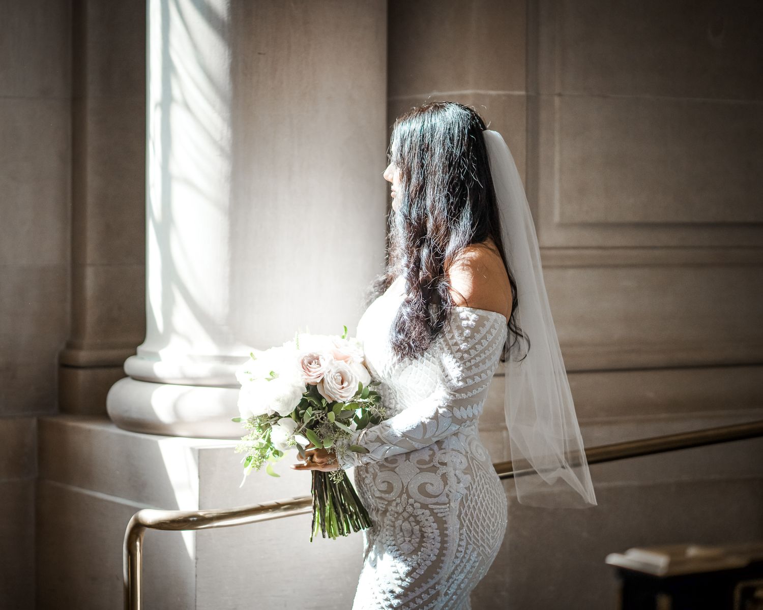 Elegant bride in lace gown holding bouquet poses by classic stone column.