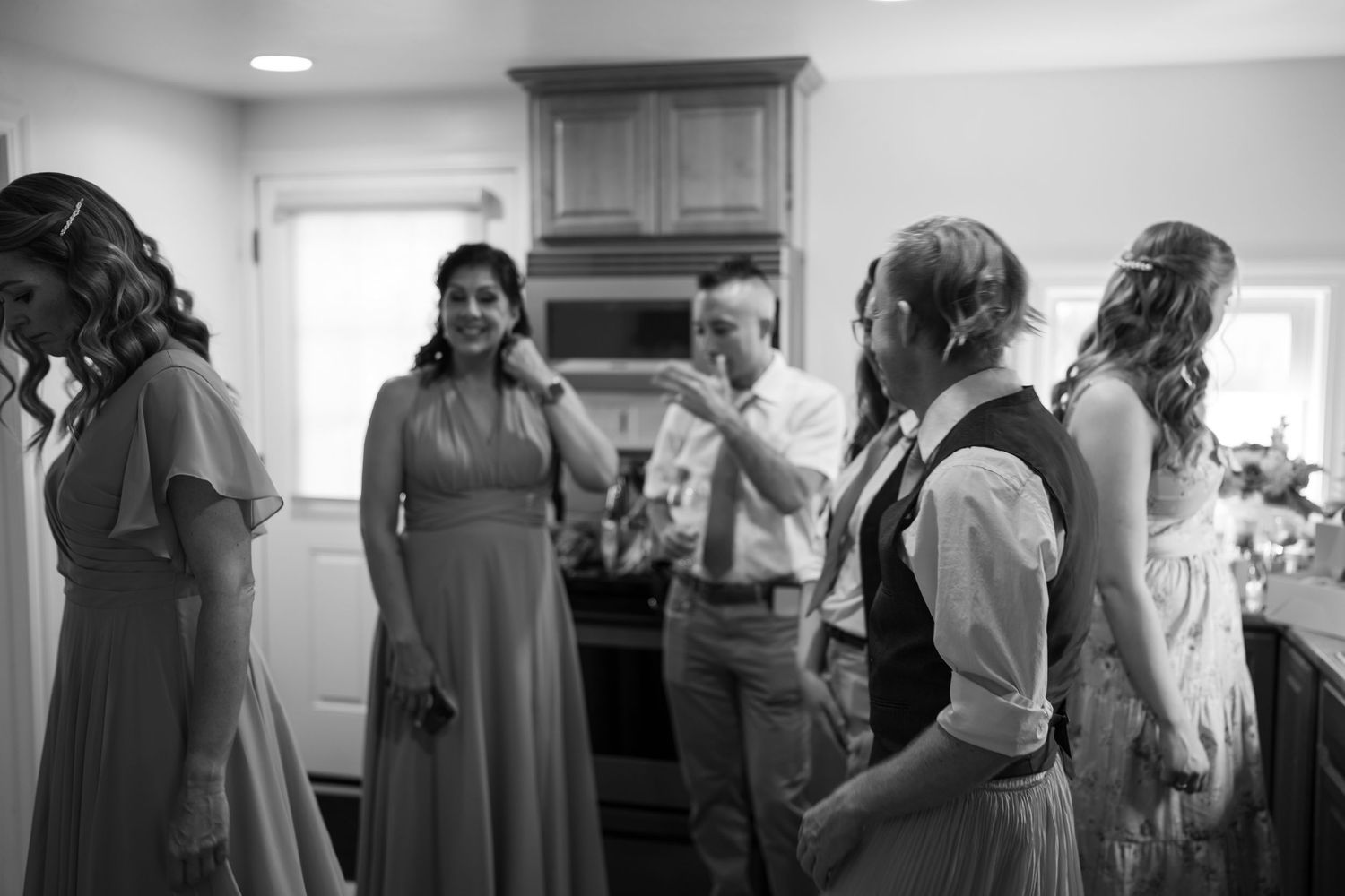 Black and white wedding photo of bridesmaids in gowns and groomsmen gathered in a kitchen during bridal party preparations.