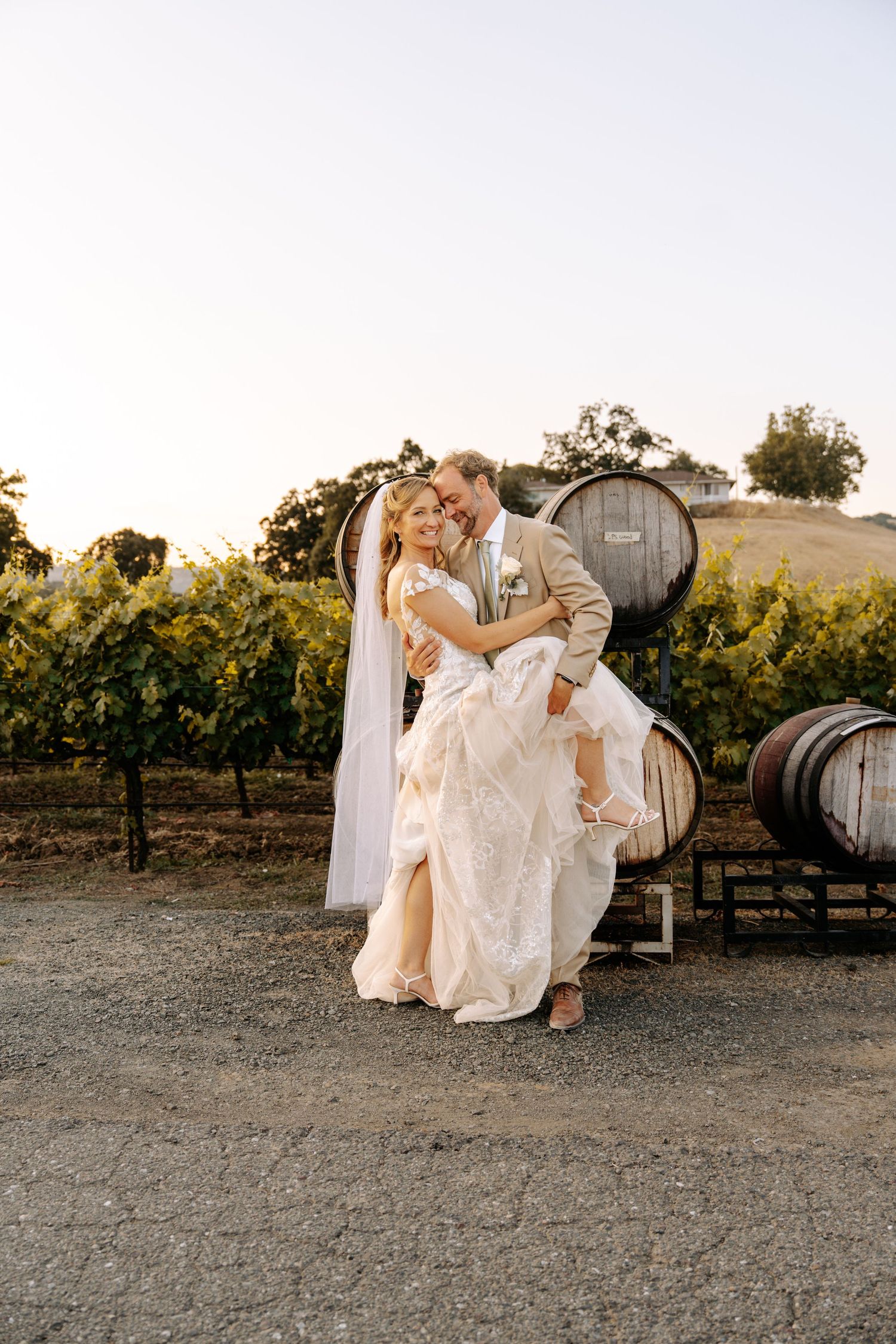 Groom lifts bride in white lace gown among vineyard wine barrels at golden hour wedding celebration.