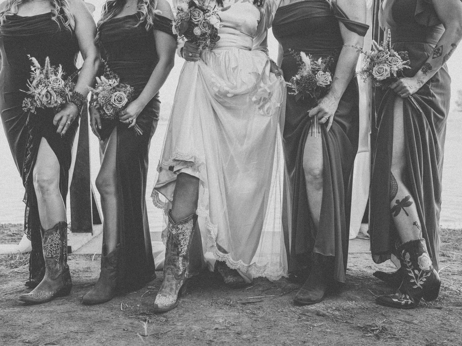 Black and white photo of bride and bridesmaids in cowboy boots lifting dresses to show footwear at rustic wedding.