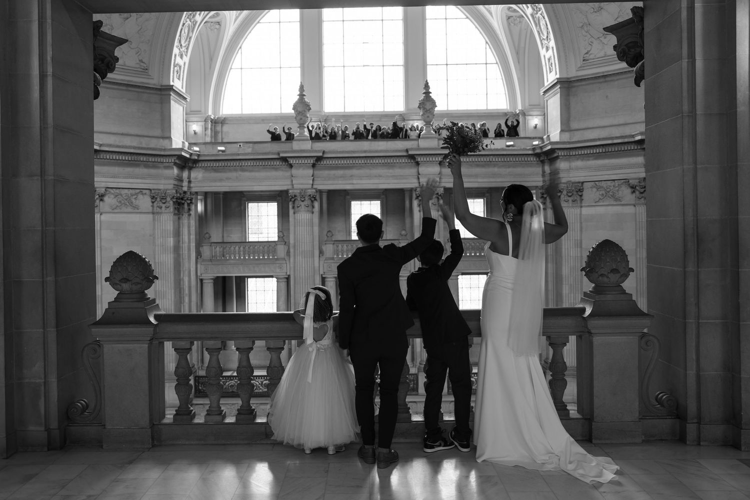 Black and white photo of a bride, groom, and flower girl celebrating at a grand wedding ceremony in an elegant hall.
