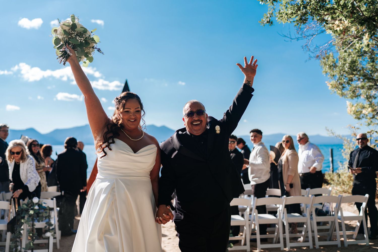 Joyful wedding celebration on waterfront with couple raising arms in celebration.