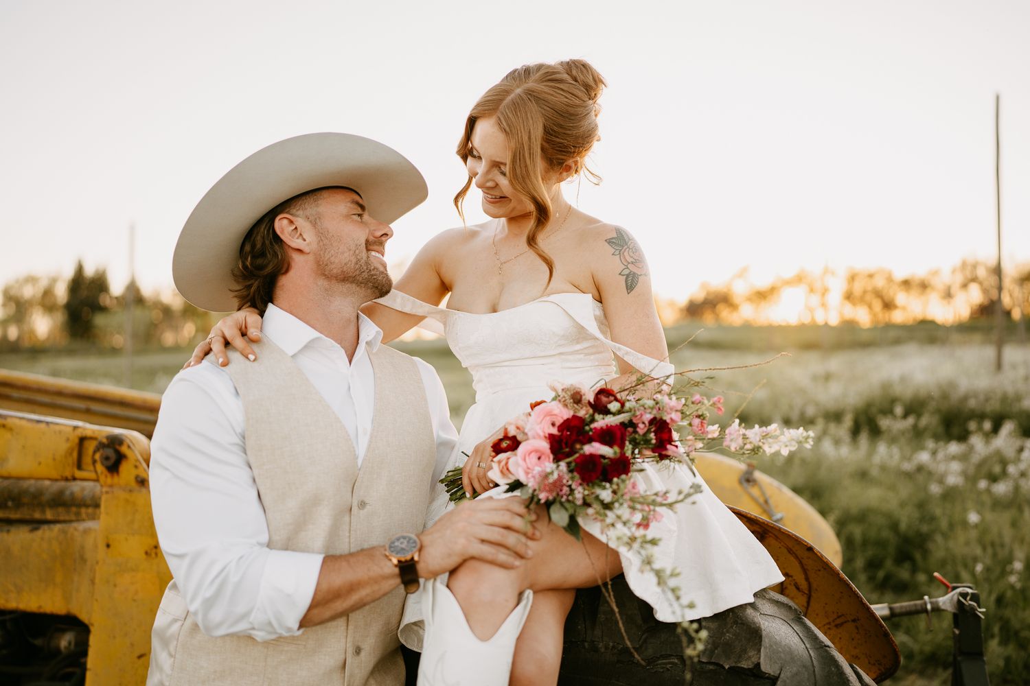 Bride and groom in rustic western wedding attire share a romantic moment outdoors at golden hour with floral bouquet.