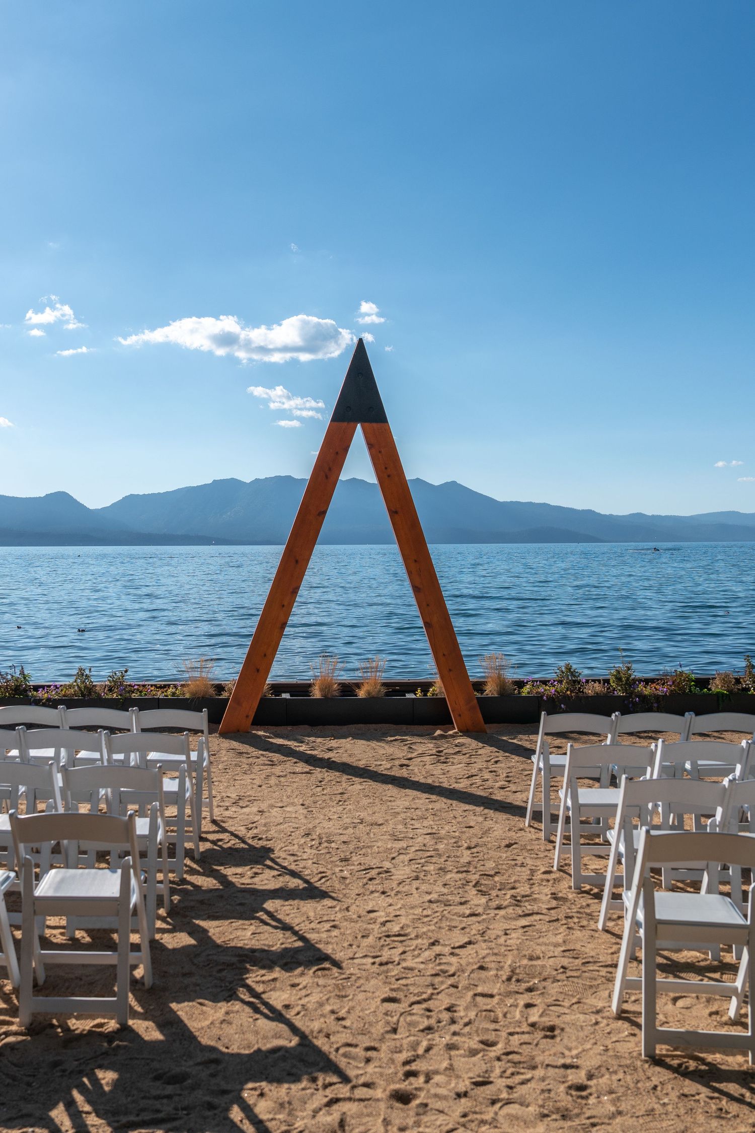 Beach wedding ceremony setup with white chairs and triangular wooden arch overlooking a mountain lake.